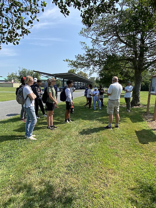 A group of students stands in front of Dr. Bill Dennison outside as he shares with them about the history of the research ponds at the lab.